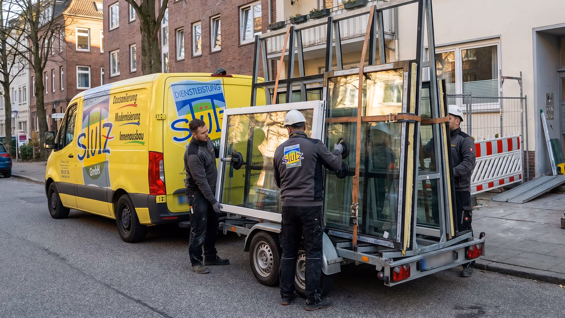 Drei Handwerker laden Fensterrahmen mit Handschuhen von einem gelben Firmenfahrzeug auf Anhänger.