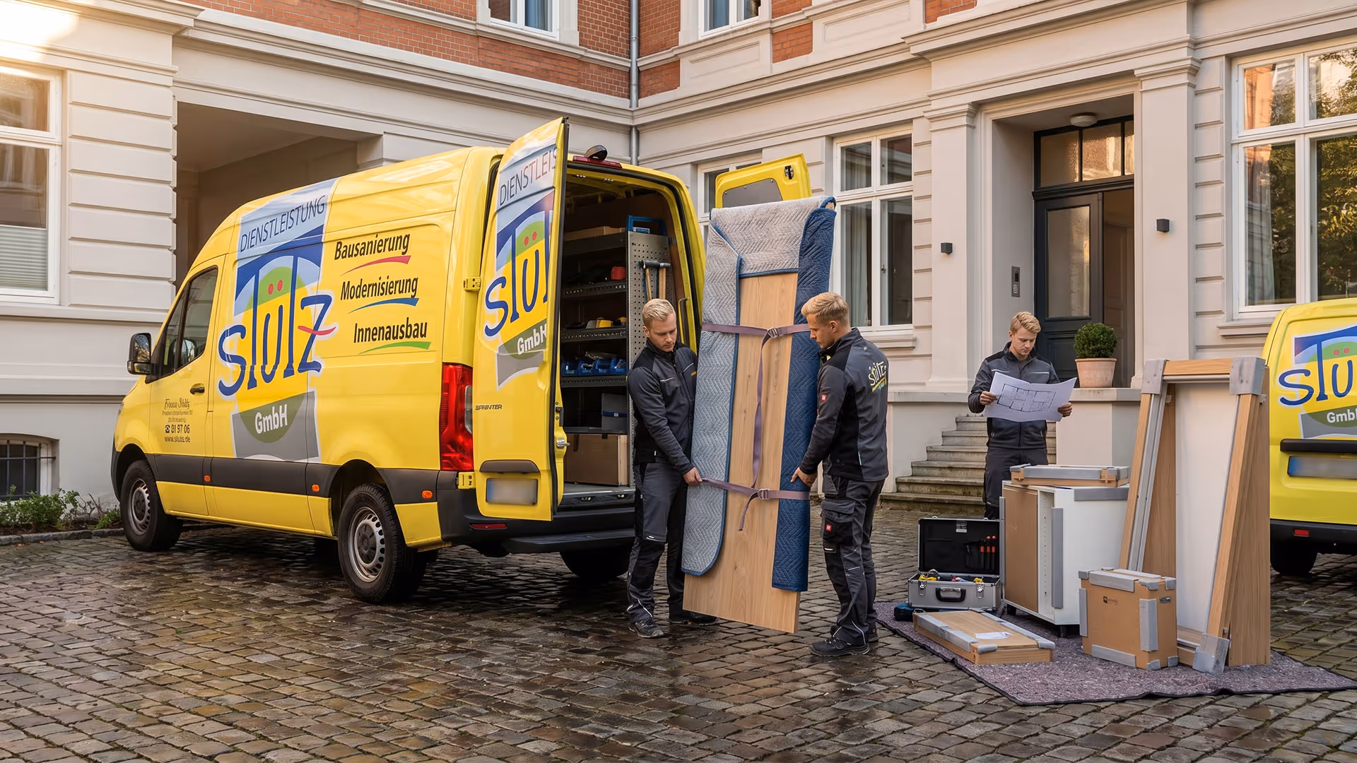 Zwei Handwerker tragen verpacktes Möbelstück aus einem gelben Transporter vor einem Gebäude.