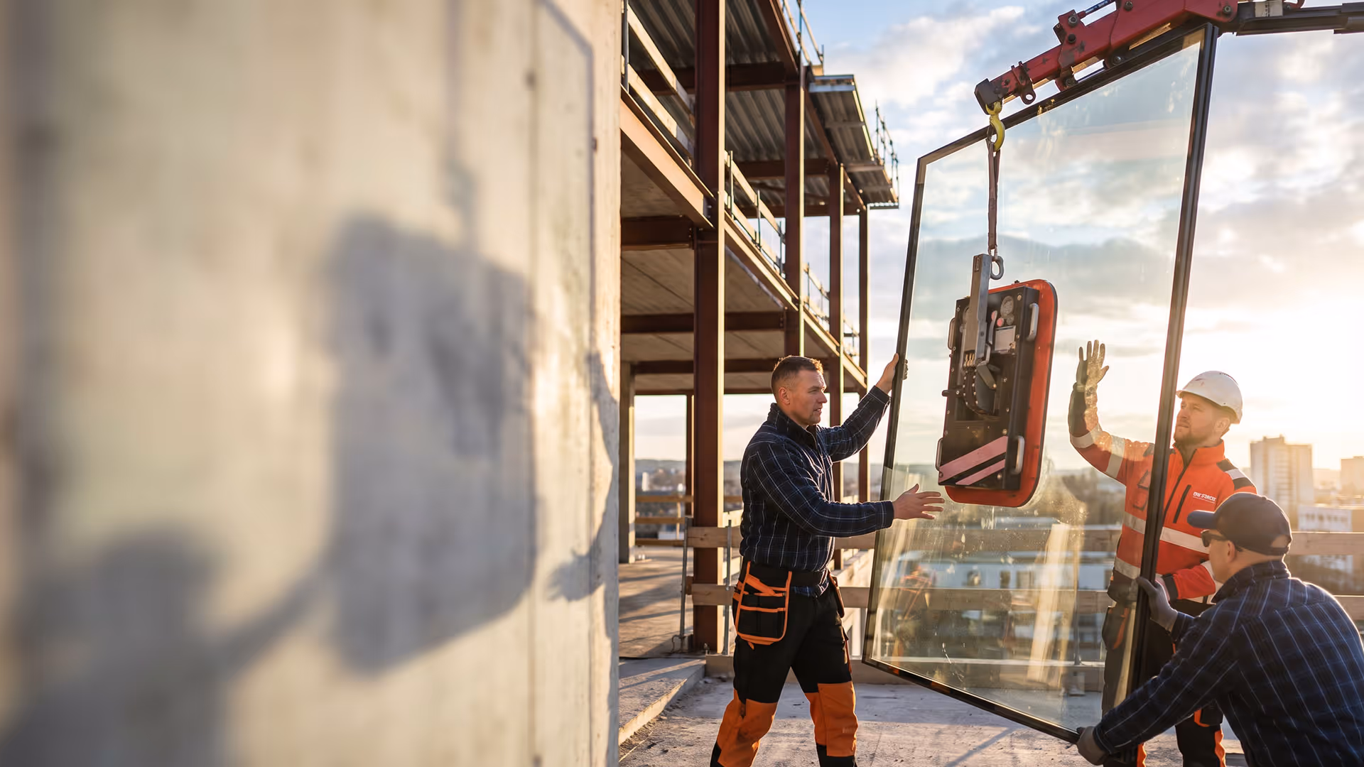 Drei Bauarbeiter installieren auf einer Baustelle mit einem Kran ein großes Glasfenster.