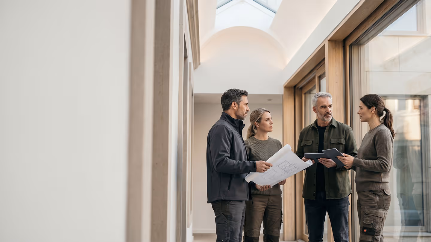 Vier Erwachsene diskutieren Pläne und Tablets in einem hellen Flur mit Fensterfront.
