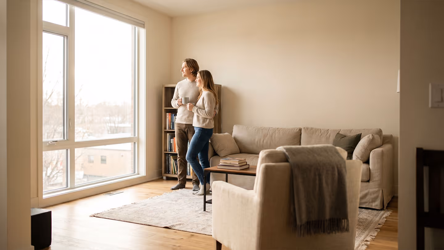 Ein Paar steht mit Tassen in der Hand und schaut durch ein großes Fenster in einem hellen Wohnzimmer.