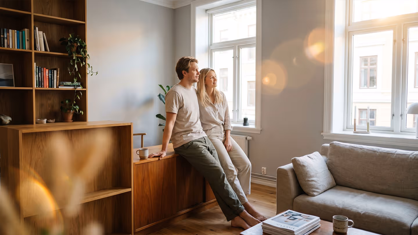 Paar sitzt entspannt auf Holzregal in hellem Wohnzimmer mit Couch, Bücherregal und großen Fenstern.