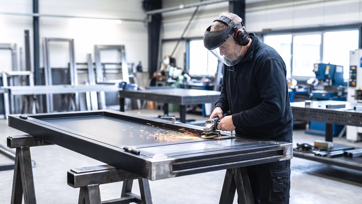 Handwerker schleift einen Metallrahmen mit Schutzhelm und Gehörschutz in einer Werkstatt.