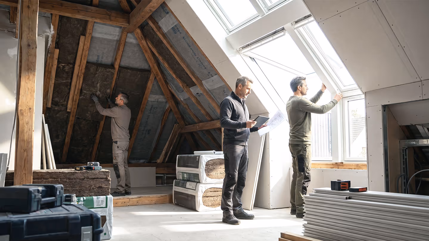 Drei Männer renovieren ein Dachgeschoss, einer bringt Dämmung an, andere prüfen Pläne und Fenster.