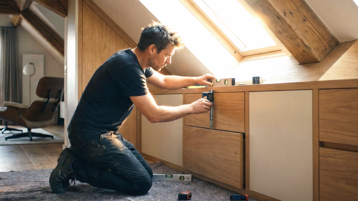 Handwerker kniet auf Teppich und misst eine Holzkommode mit Wasserwaage im Dachzimmer.