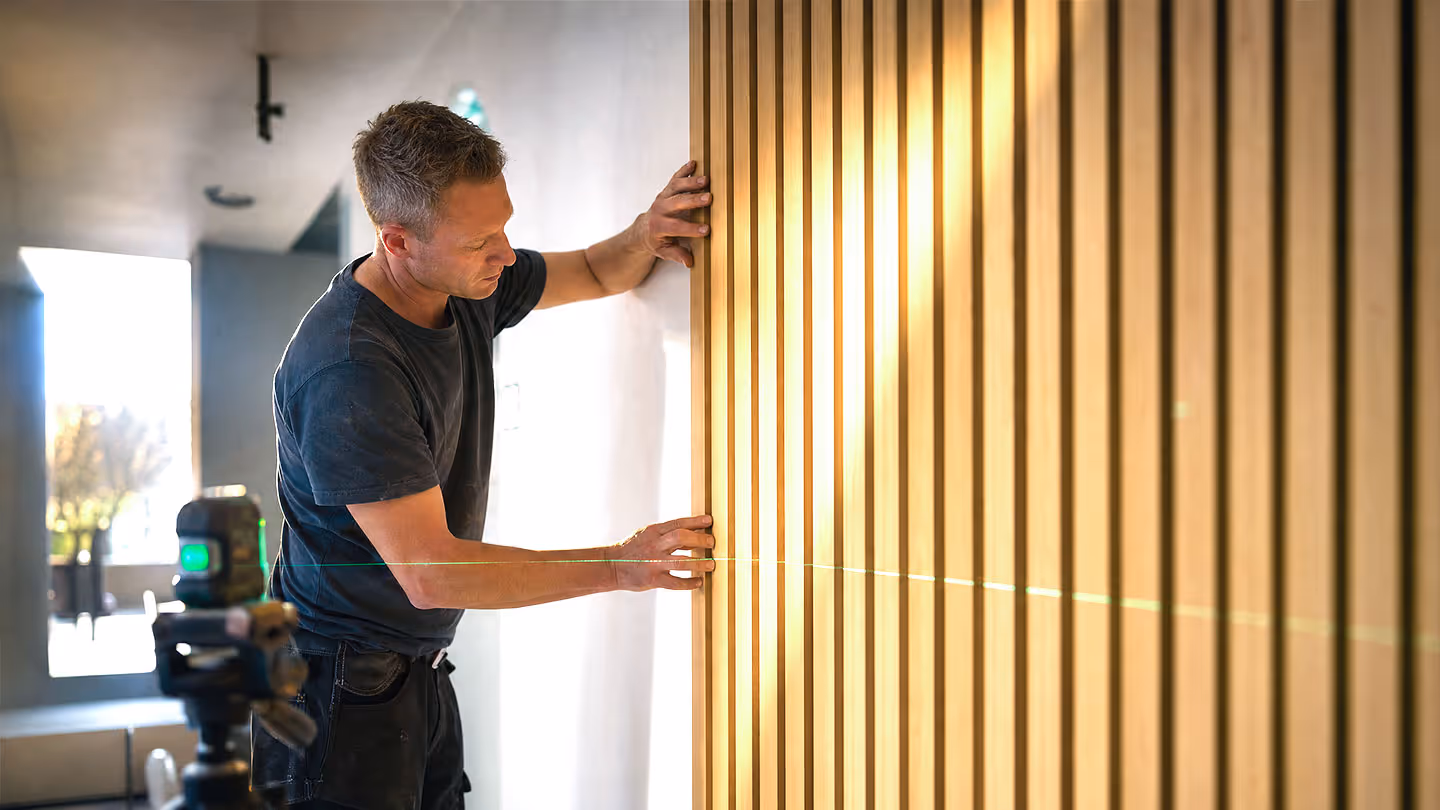 Mann prüft mit Laser-Wasserwaage vertikale Holzpaneele an Wand in modernem Raum.