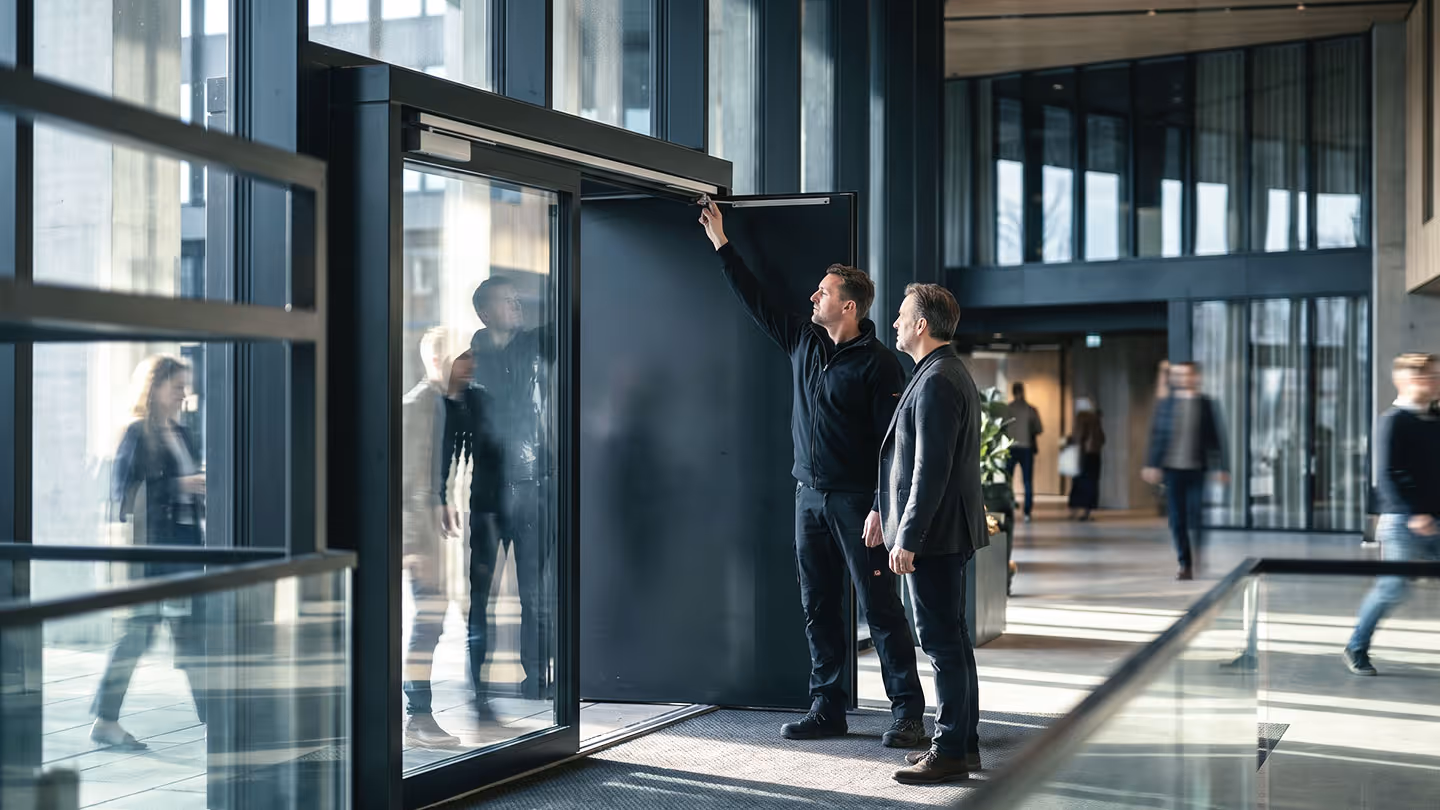 Zwei Männer überprüfen eine Tür in einem modernen Gebäude mit großen Glasfenstern.