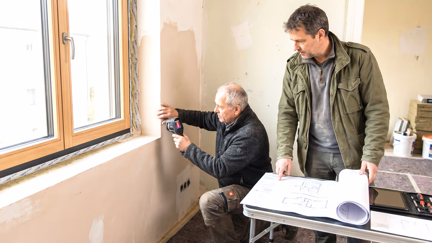Zwei Handwerker prüfen mit Wärmebildkamera und Bauplänen einen Fensterrahmen während der Modernisierung.
