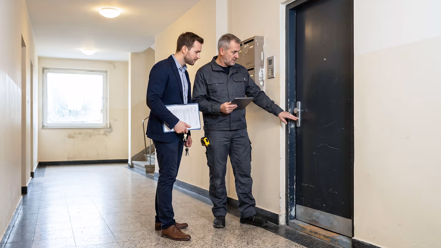Zwei Männer stehen im Flur vor einer schwarzen Tür, einer hält Schlüssel und Aktenordner.