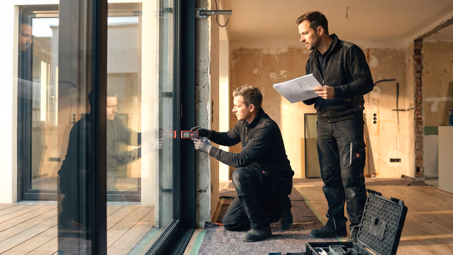 Zwei Handwerker messen ein Fenster mit Lasergerät, einer hält Pläne in einem Rohbauzimmer.