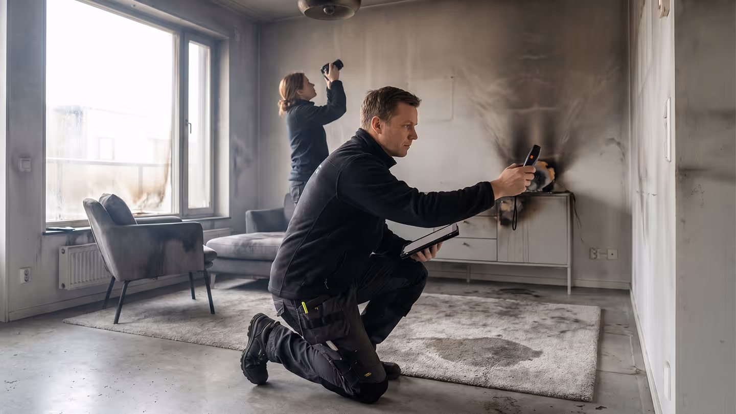 Zwei Personen inspizieren und fotografieren Brandschäden in einem verrauchten Wohnzimmer.
