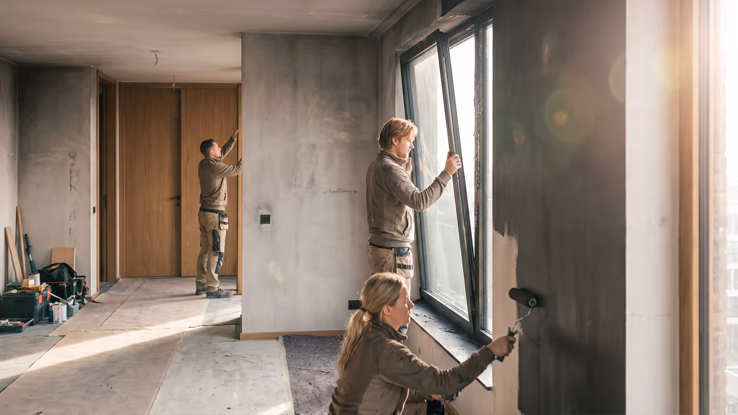 Drei Handwerker renovieren eine Wohnung mit Fensterinstallation, Wandbemalung und Türprüfung.