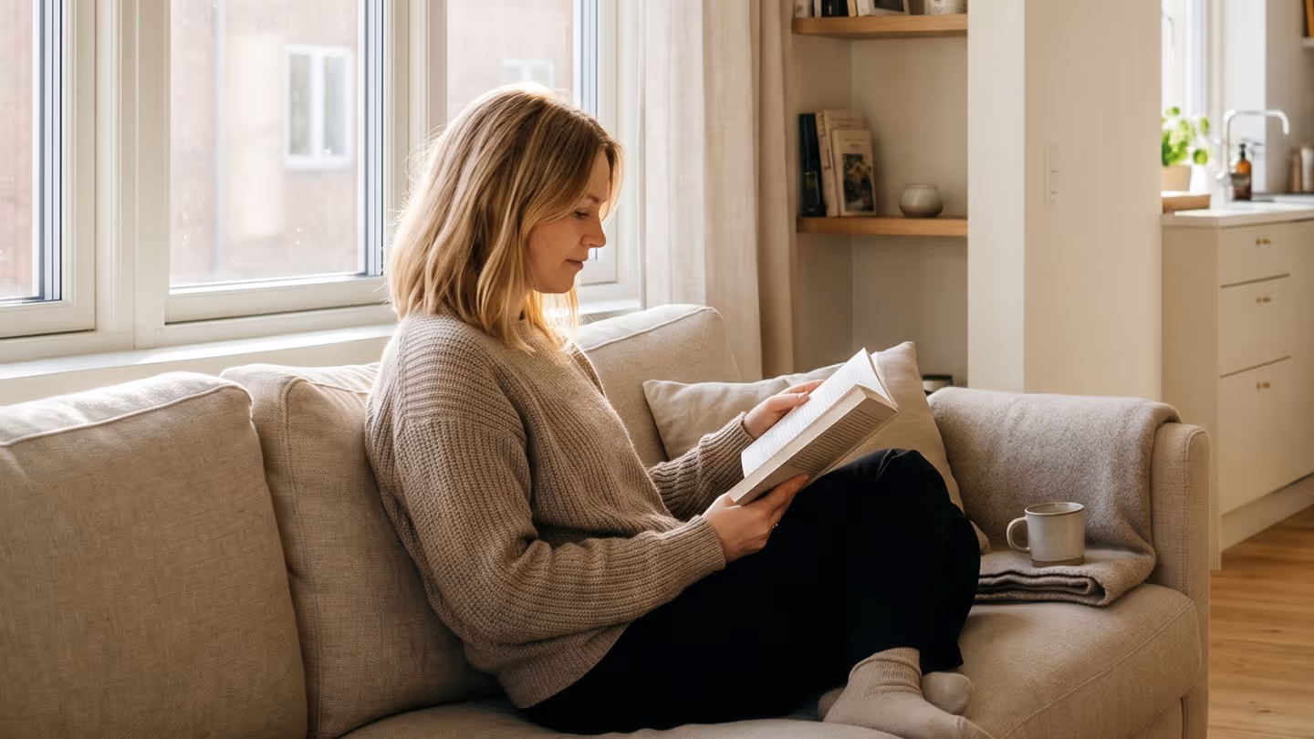 Frau sitzt auf Sofa und liest Buch in hellem Wohnzimmer mit Kaffeetasse neben ihr.