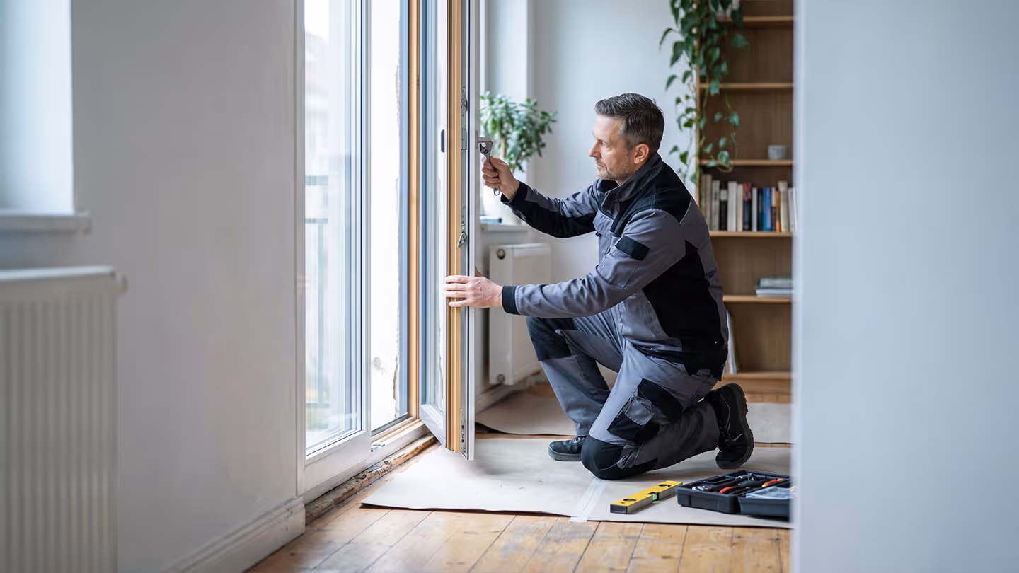 Handwerker kniet und justiert ein großes Fenster mit Werkzeug in einem hellen Raum.