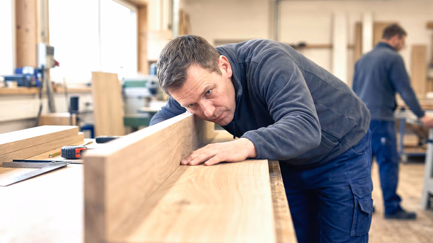 Tischler prüft sorgfältig eine Holzplatte in einer Werkstatt voller Holzmaterialien und Werkzeug.