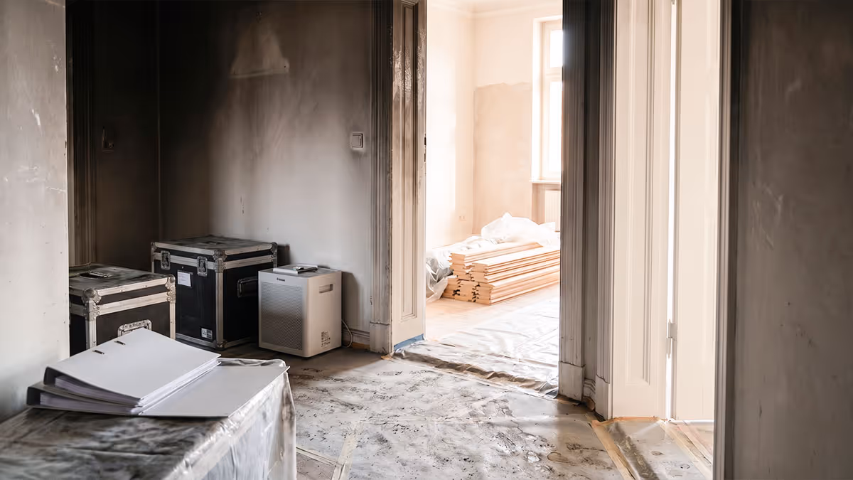 Rauchgeschädigter Innenraum mit staubbedecktem Boden, Kisten und Holzbrettern in hellem Raum dahinter.